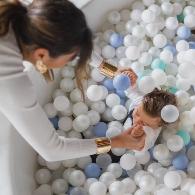 Overhead view of an adult holding a toddler's hand in the white ball pit filled with white, pearl, blue and mint balls