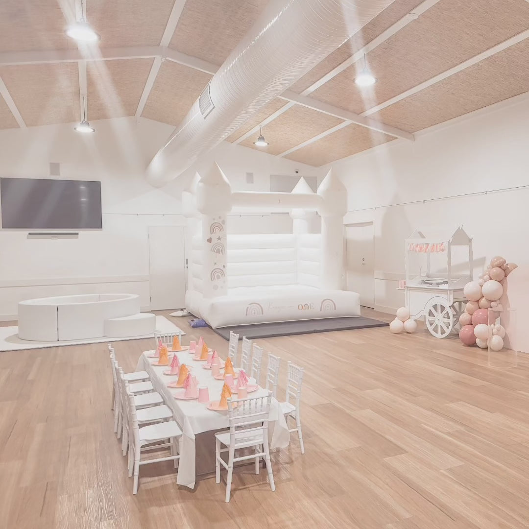 White bouncy castle with white and gold balloon garland in a marquee with festoon lights, grey mats and a timber table in front