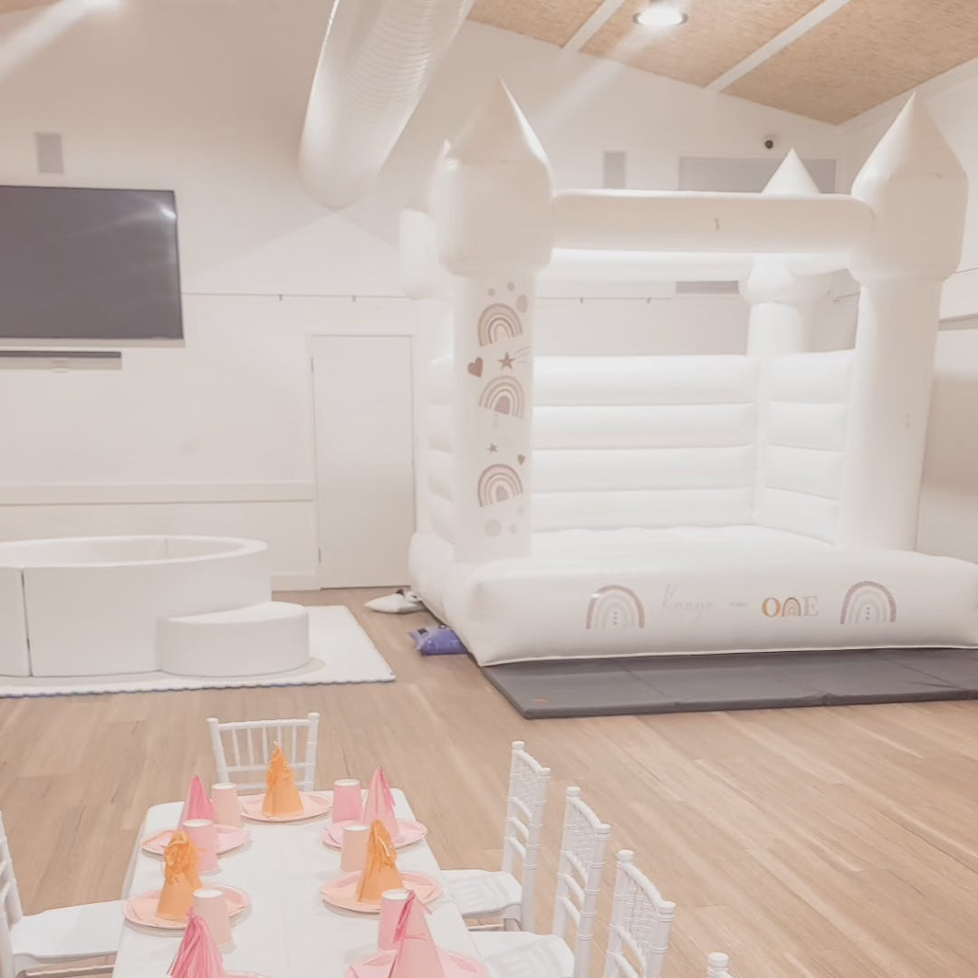 White castle with boho rainbow decals and a round white ball pit in an indoor hall, kids table with pink and orange party hats
