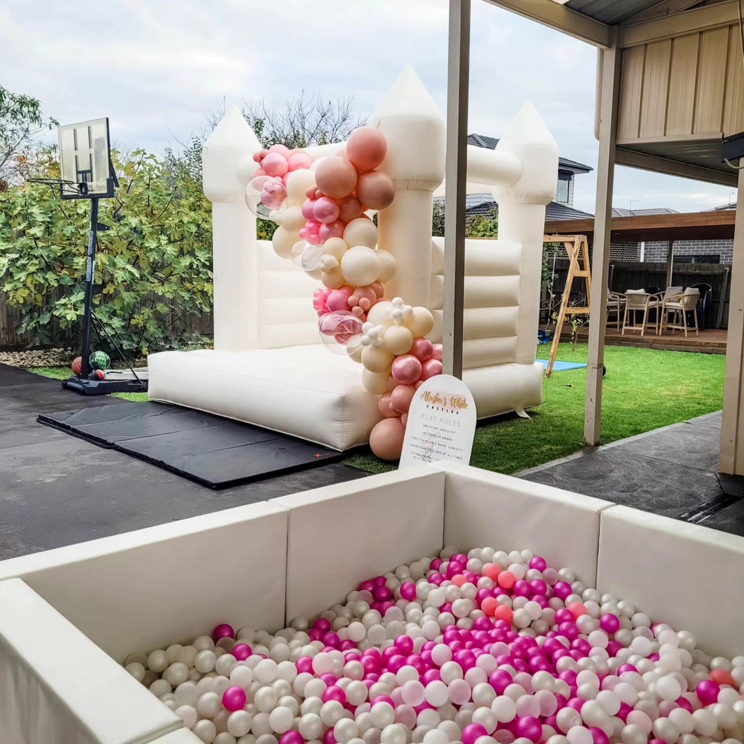 Hot pink, white and blush balls in a white ball pit in front of a white bouncy castle with a rose gold garland in a backyard