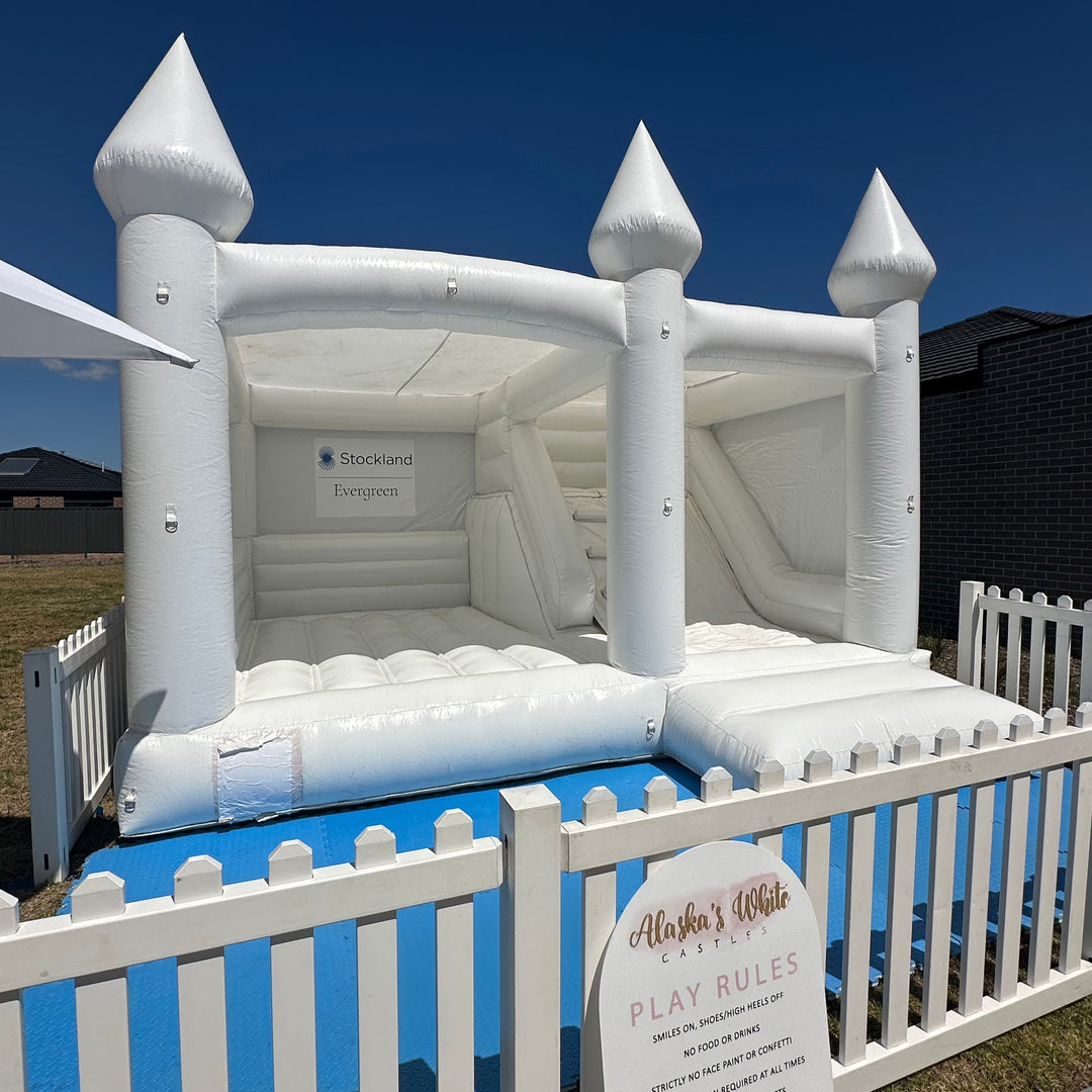 William white castle with slide inside white picket fencing at a Stockland Evergreen community event with a play rules sign