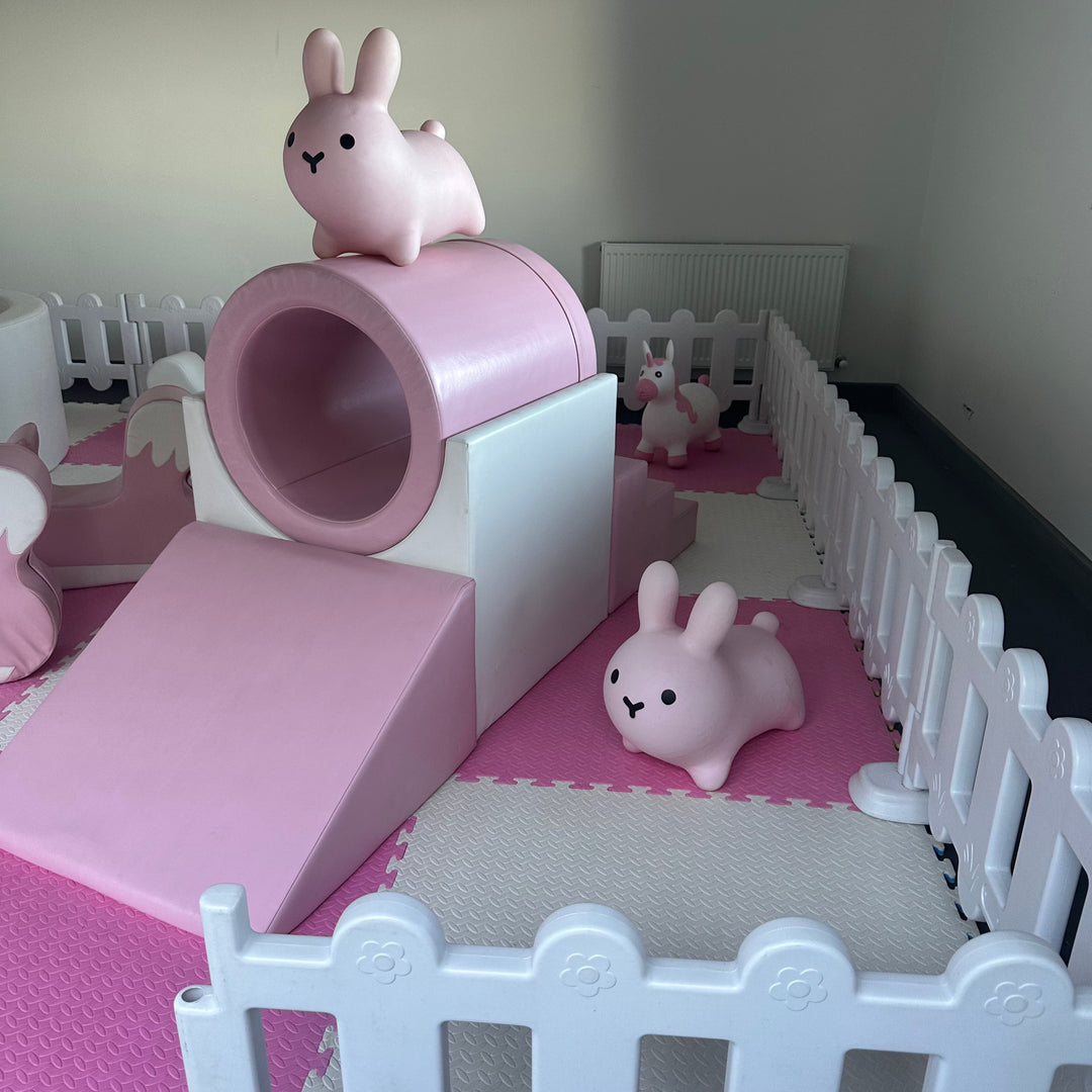 Pink tunnel and slide with a bunny hopper on top and another bunny and unicorn hopper on the mats inside a white picket fence