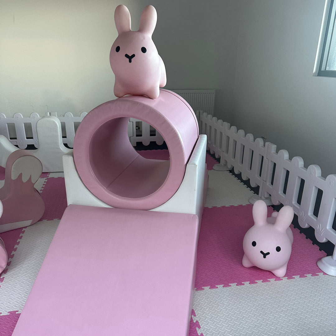 Close-up of the pink tunnel and slide with a bunny hopper perched on top and a second bunny hopper on the pink and white mats