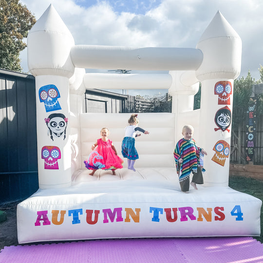 Dia de Los Muertos sugar skull decals on a white bouncy castle with kids bouncing and an Autumn turns 4 decal on the front