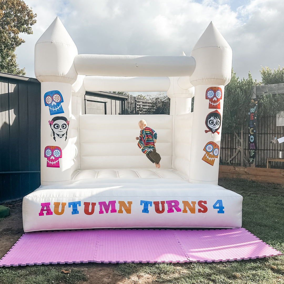 White bouncy castle with colourful Dia de Los Muertos skull decals on the pillars, a child mid-jump and purple mats on grass