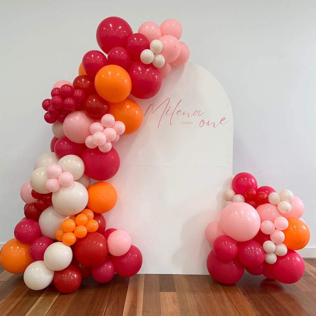 White arch backdrop with a pink script decal reading Milena turns one and a hot pink, orange and blush balloon garland