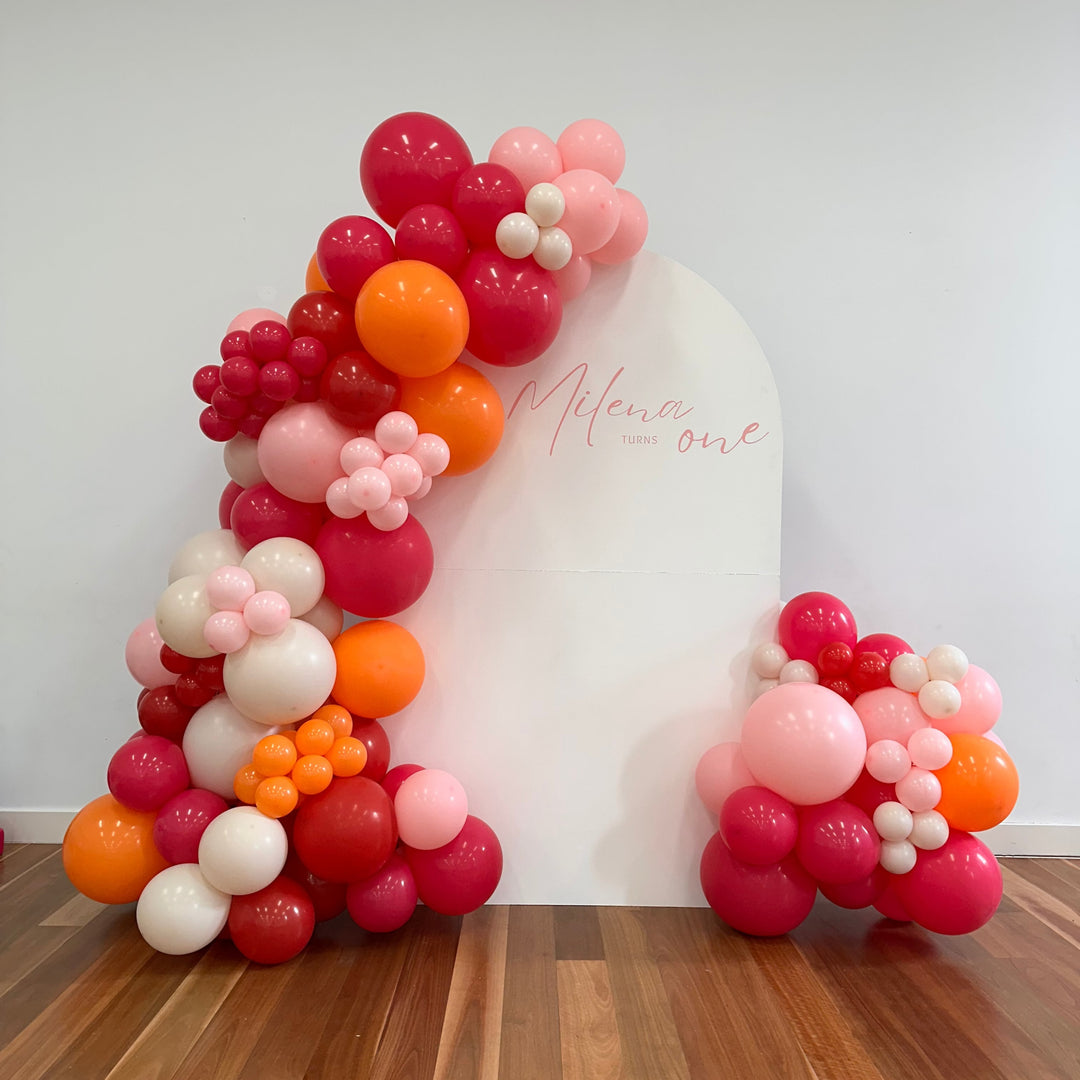 White arch backdrop with a red, orange, pink and white balloon garland and a decal reading Milena turns one on a timber floor