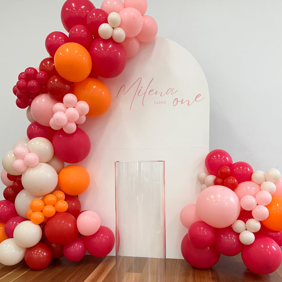 Milena hot pink, red, orange and blush balloon garland on a white arch backdrop with a pink Milena turns one decal and plinth