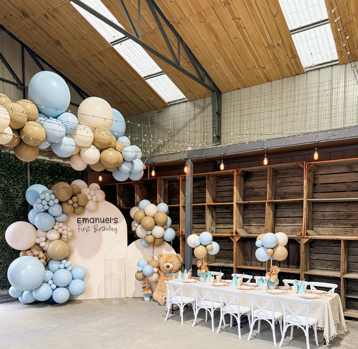 Emanuel's First Birthday arch backdrop with a blue and tan balloon garland, teddy bears and a kids table in a timber barn