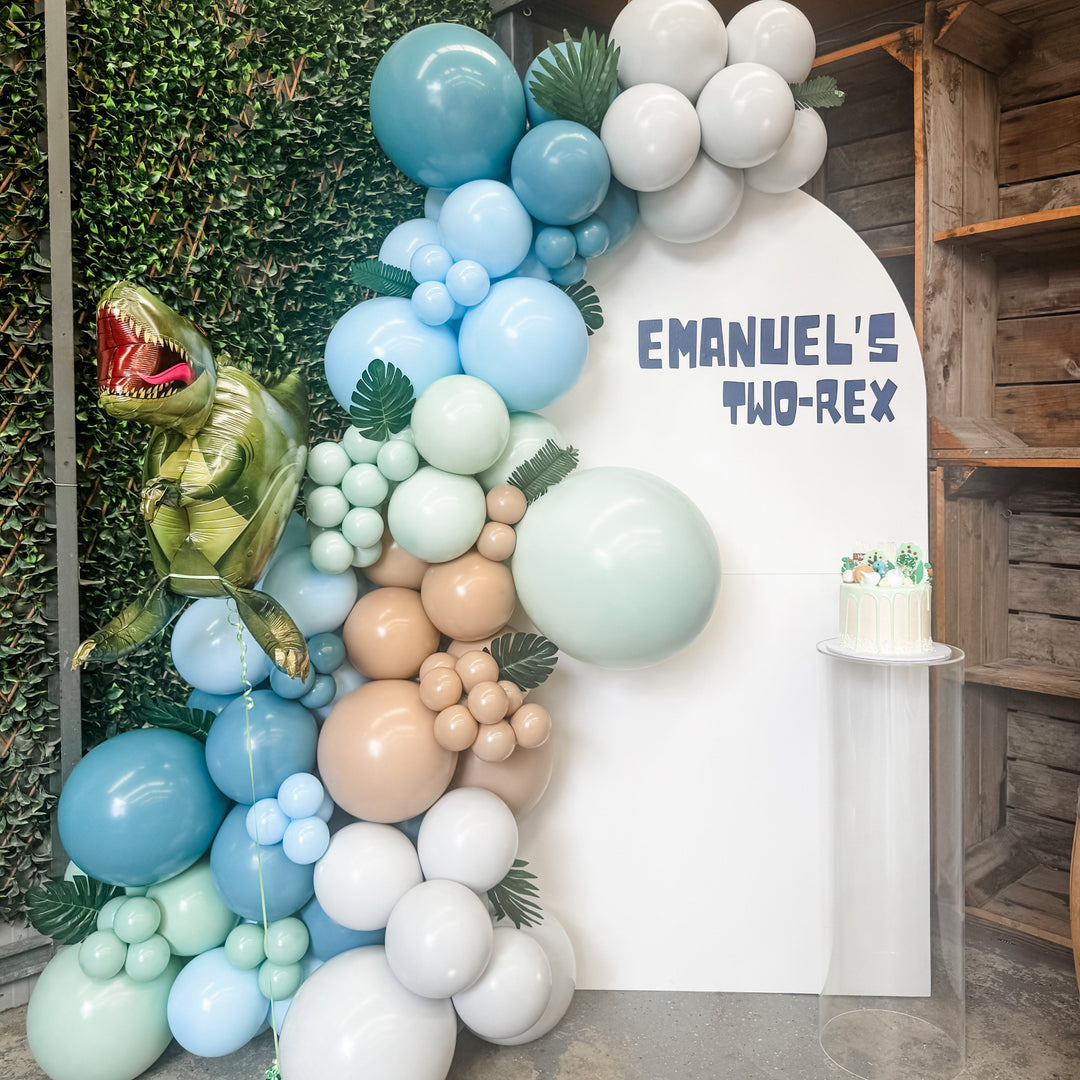 White arch backdrop with an Emanuel's Two-Rex decal, a blue, sage and tan dinosaur balloon garland and a cake on a plinth