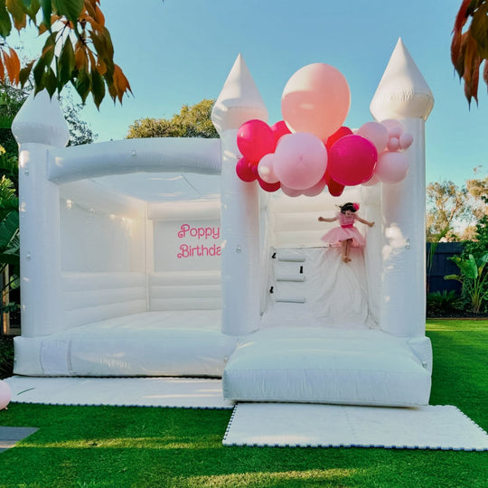William white bouncy castle with slide, pink balloon garland and Poppy's Birthday decal on a lawn for party hire in Melbourne