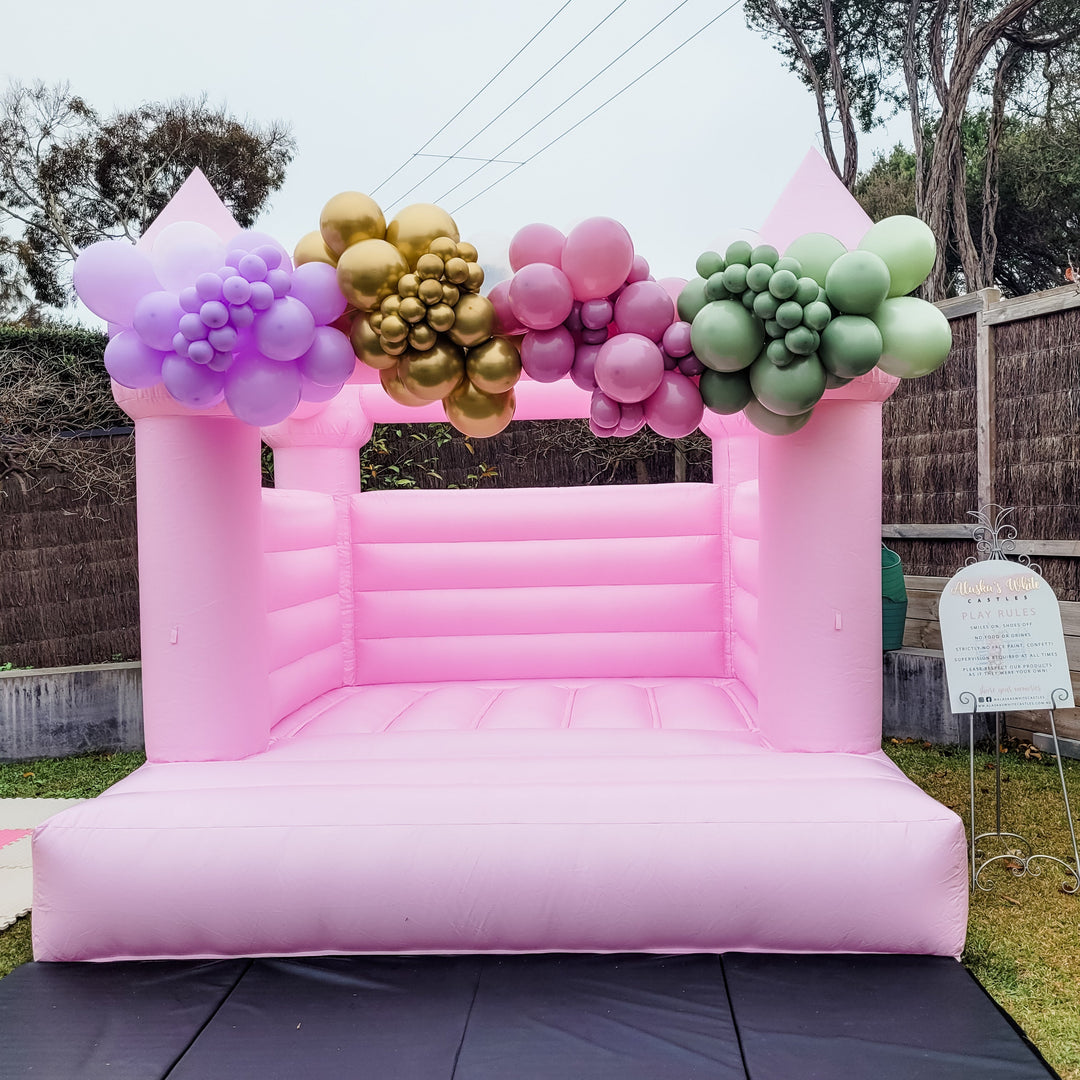 Pink bouncy castle with lilac, gold, mauve and sage green balloon clusters along the top beam and black crash mats in front
