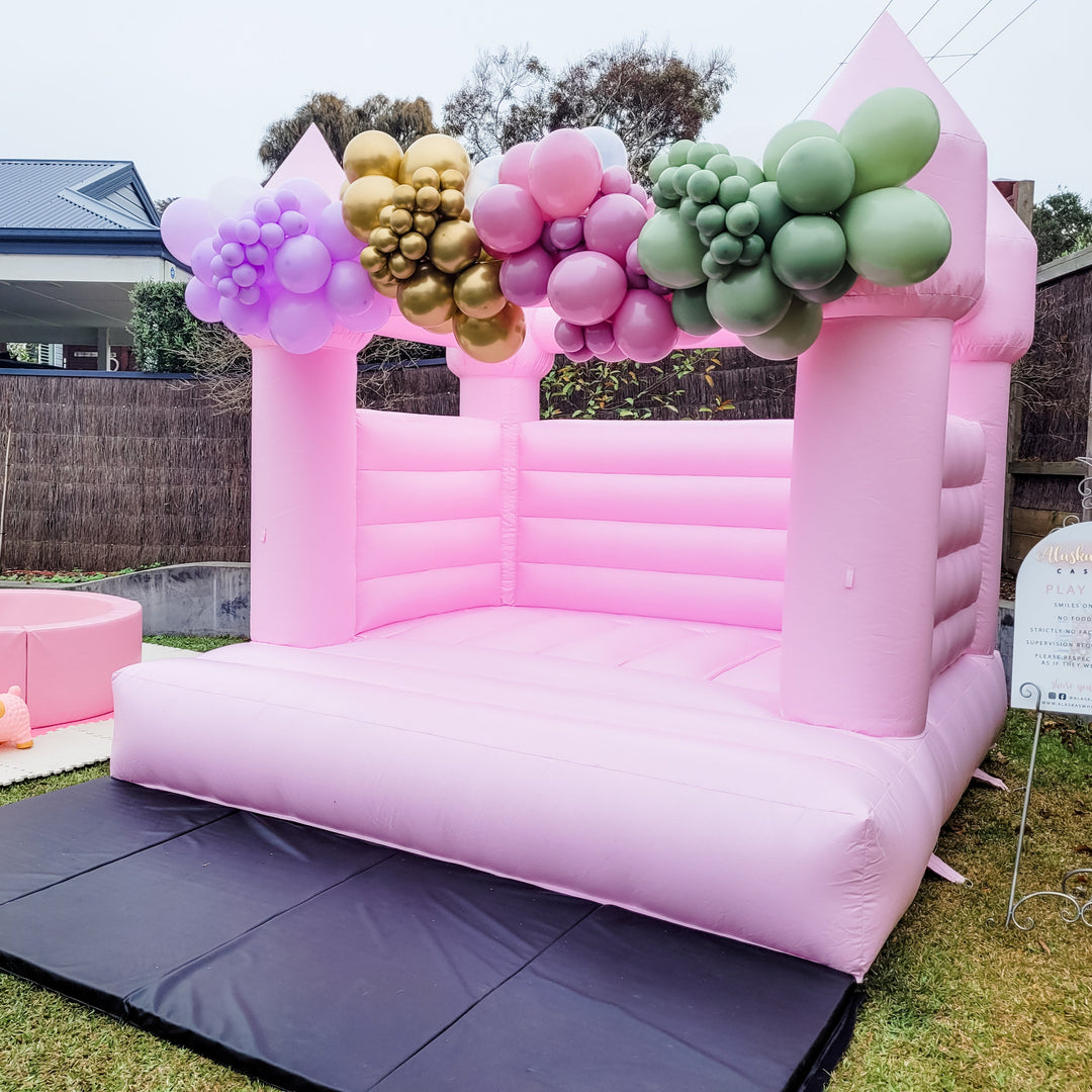 Angled view of the pastel pink bouncy castle with lilac, gold, mauve and sage balloon clusters on top and a play rules sign