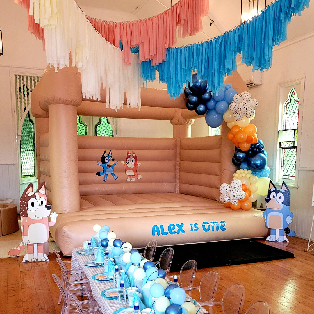 Tan bouncy castle in a hall with Bluey and Bingo decals, an Alex is One front decal and a blue and orange balloon garland