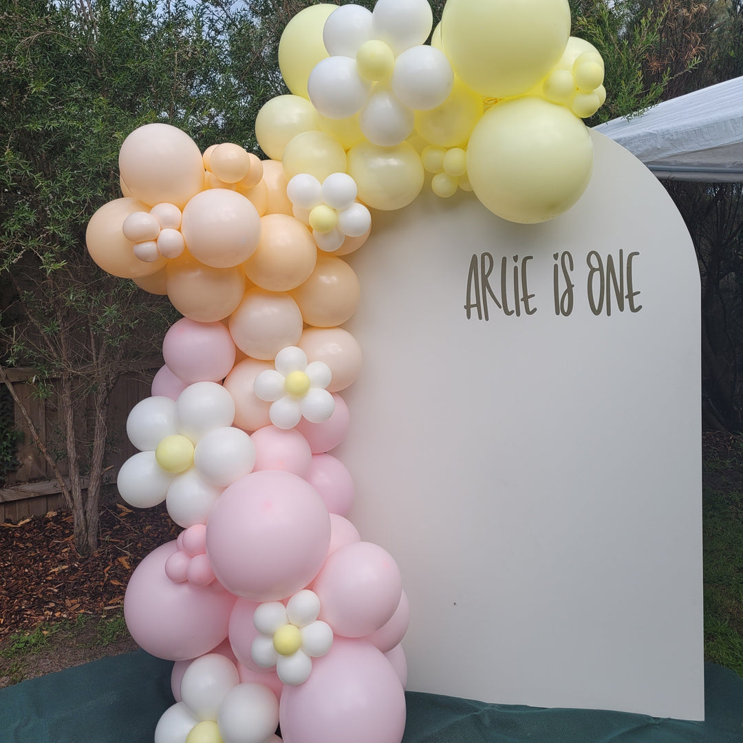 Arlie balloon garland of pastel pink, peach and yellow balloons with daisy accents on a cream arch beside a marquee
