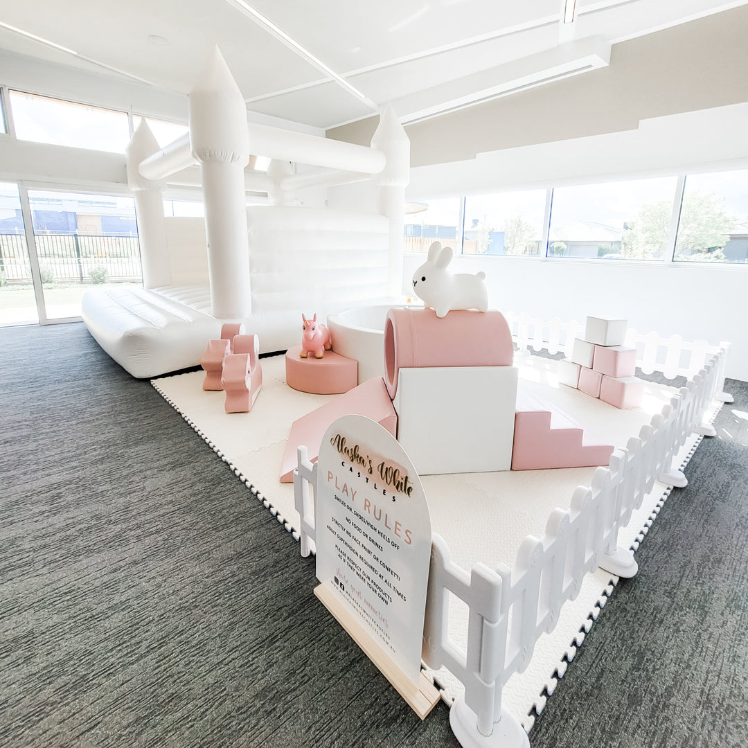 Pale pink tunnel, slide, steps and ball pit with bunny and unicorn hoppers in front of a white bouncy castle in an indoor office