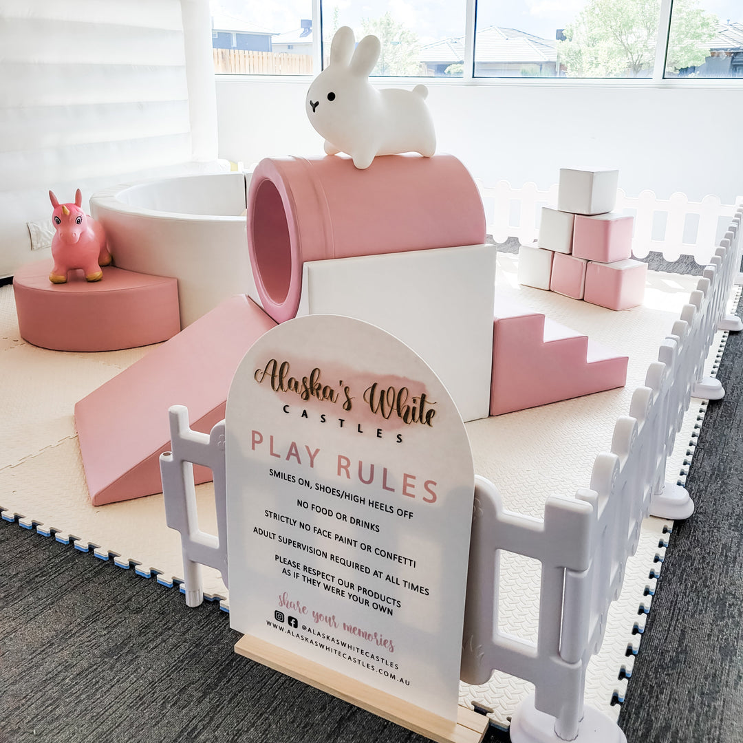 Close-up of the play rules sign beside a pink tunnel with a white bunny hopper on top and a unicorn hopper and slide behind