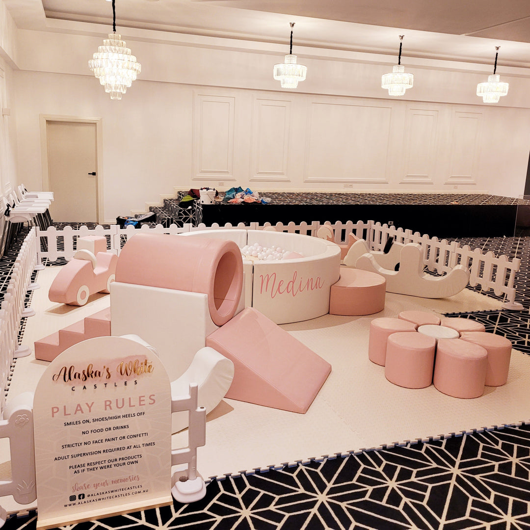 Baby pink ball pit reading Medina, ring tunnel, slide and flower seat beside a Play Rules sign in a ballroom with chandeliers