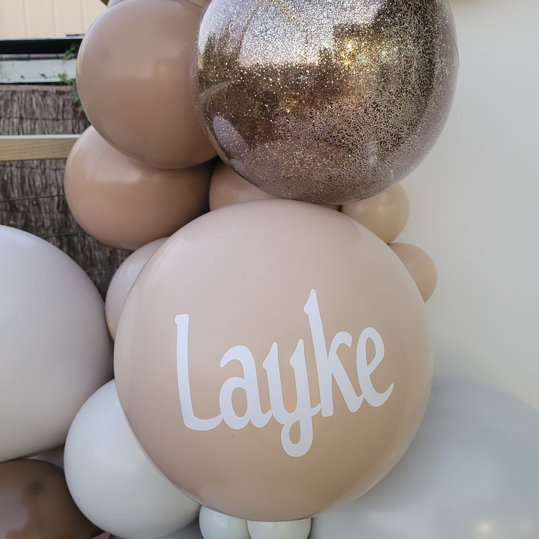 Close-up of a custom balloon decal reading Layke in white on a tan balloon beside a gold glitter orb at a Melbourne party