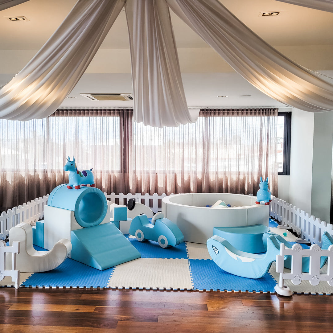 Wide view of the baby blue soft play fenced with white pickets under ceiling drapes, unicorn hoppers on the tunnel and ball pit