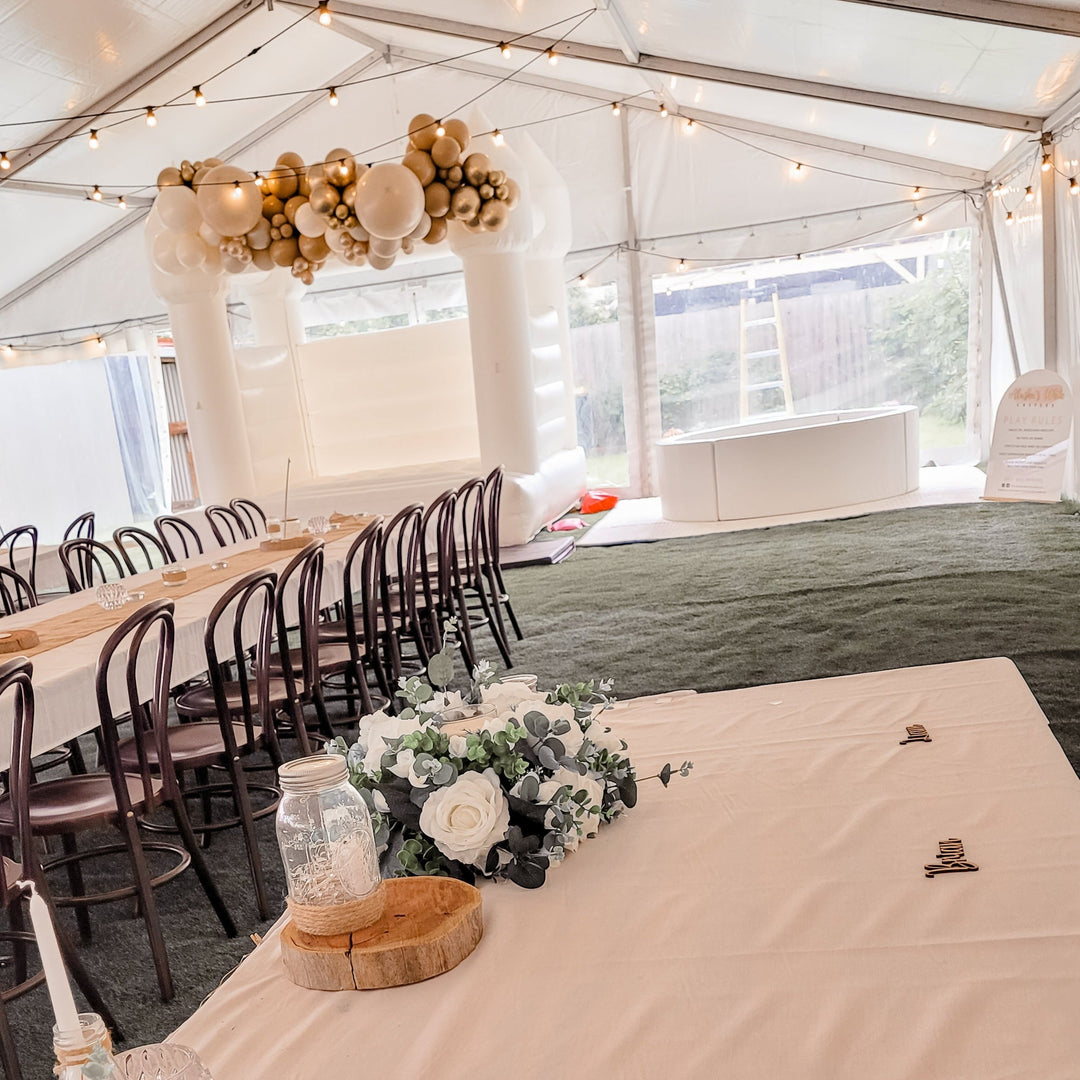 Marquee party with long tables, bentwood chairs and white florals and a white bouncy castle with gold garland behind