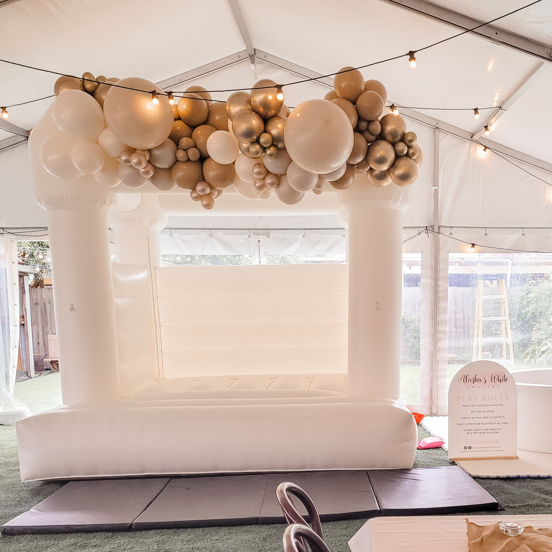 White bouncy castle topped with a white, tan and gold balloon garland in a marquee with festoon lights and a play rules sign
