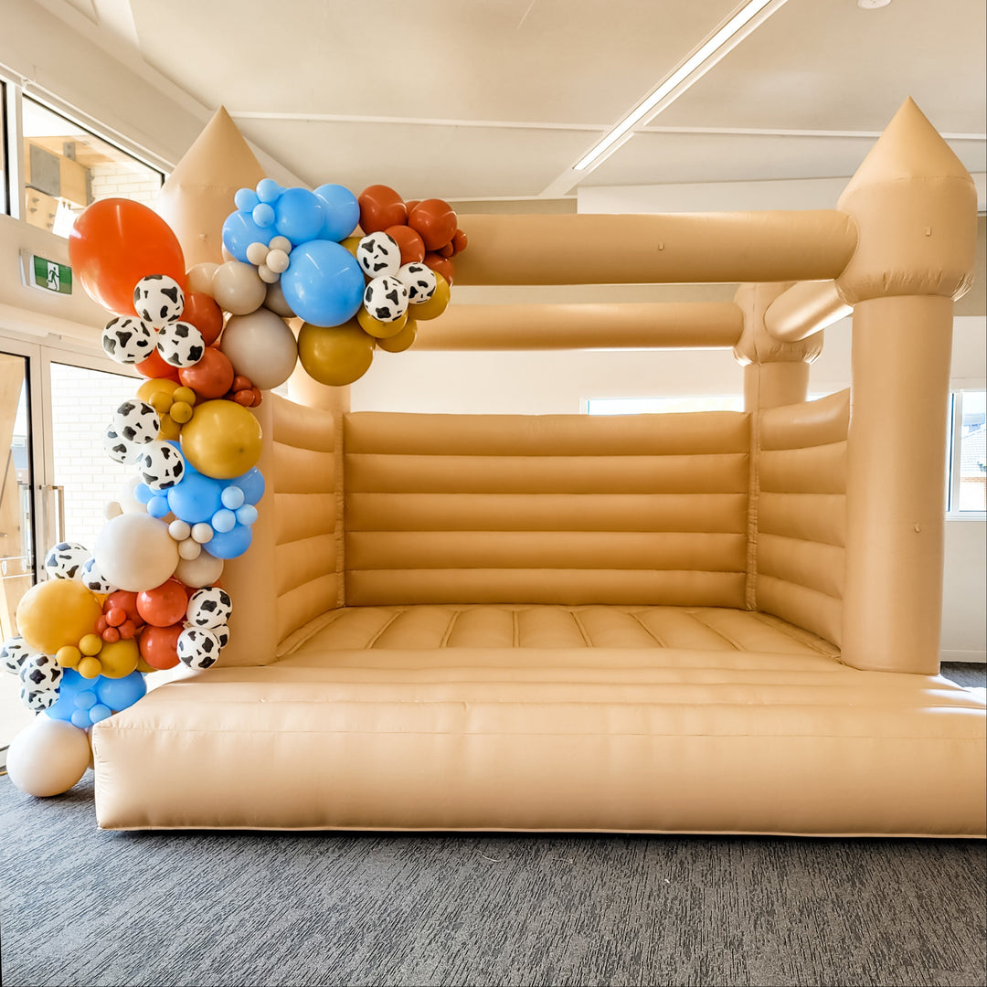 Front view of a tan bouncy castle with the Woody balloon garland in blue, rust, mustard and cow print down the left pillar