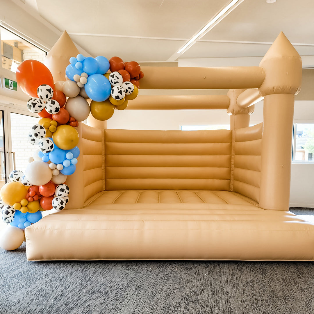 Tan bouncy castle indoors with a rust, blue, mustard and cow print balloon garland on the front tower in a function room