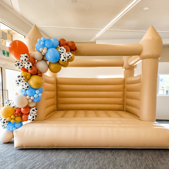 Woody balloon garland in rust, mustard, blue and cow print draped on a tan bouncy castle indoors, for party hire in Melbourne
