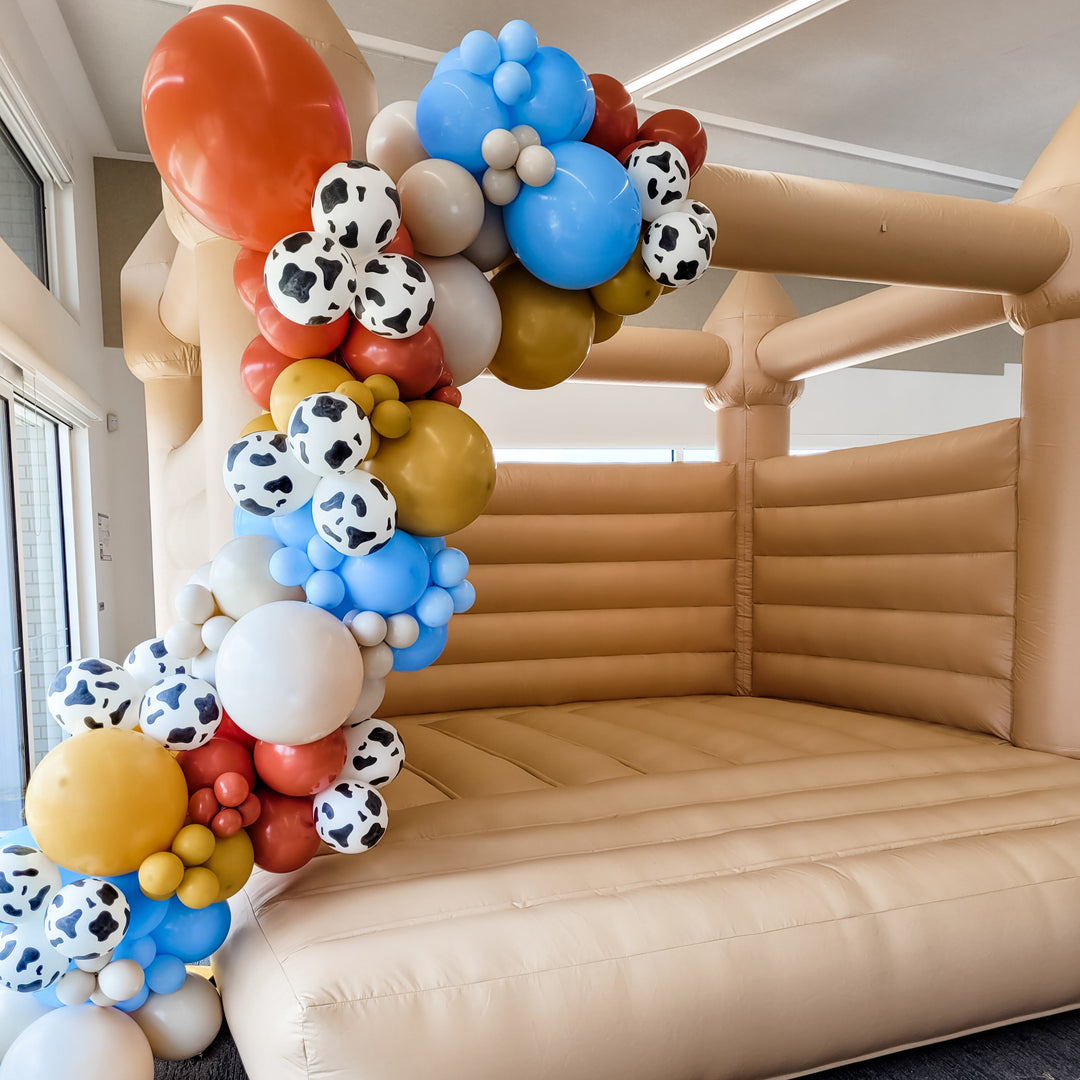 Close-up of the Woody balloon garland with cow print, sky blue, rust, mustard and grey balloons on a tan castle corner