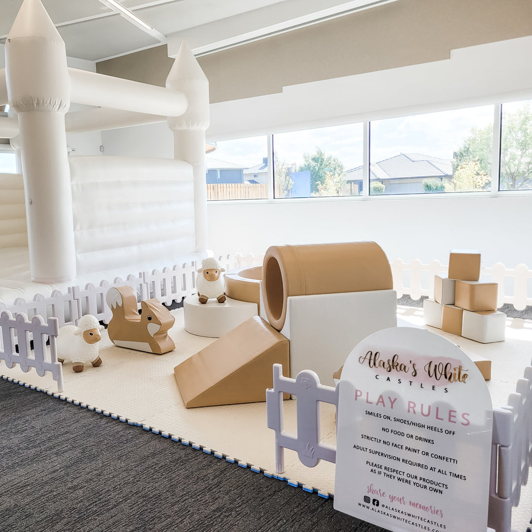 Tan tunnel slide, fox rocker and two sheep hoppers on cream mats beside a white bouncy castle inside a white picket fence