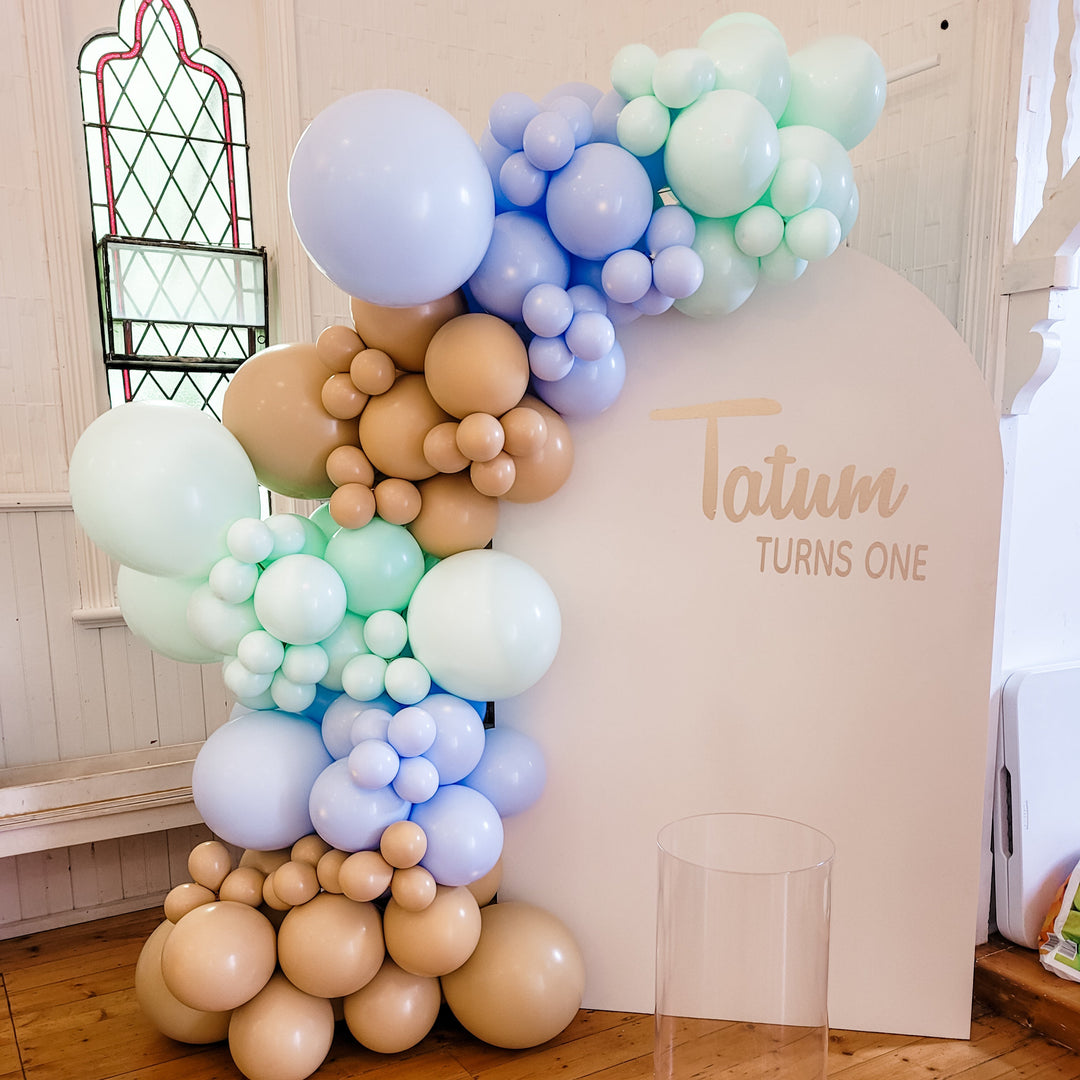 Tatum balloon garland in lilac, mint, pale blue and tan on a cream arch reading Tatum turns one for party hire in Melbourne
