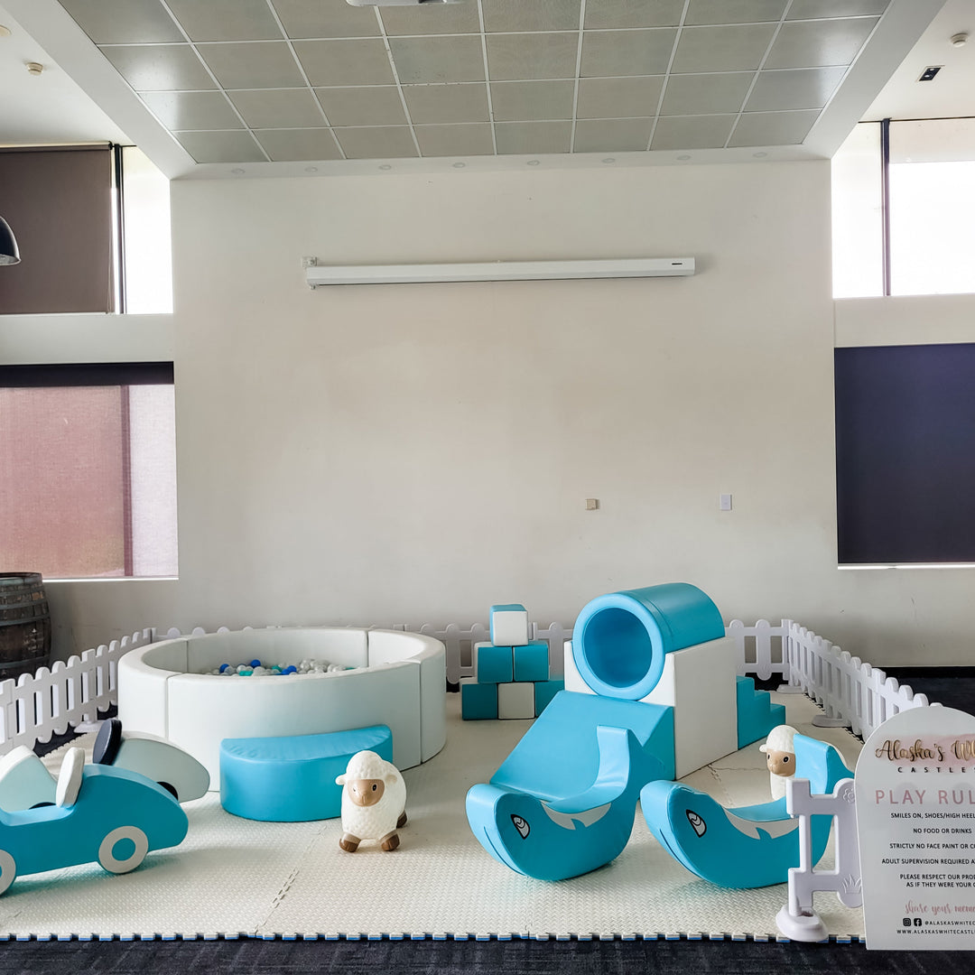 Full baby blue soft play set in a white picket fence in a plain function room with a ball pit, tunnel slide, car and rockers