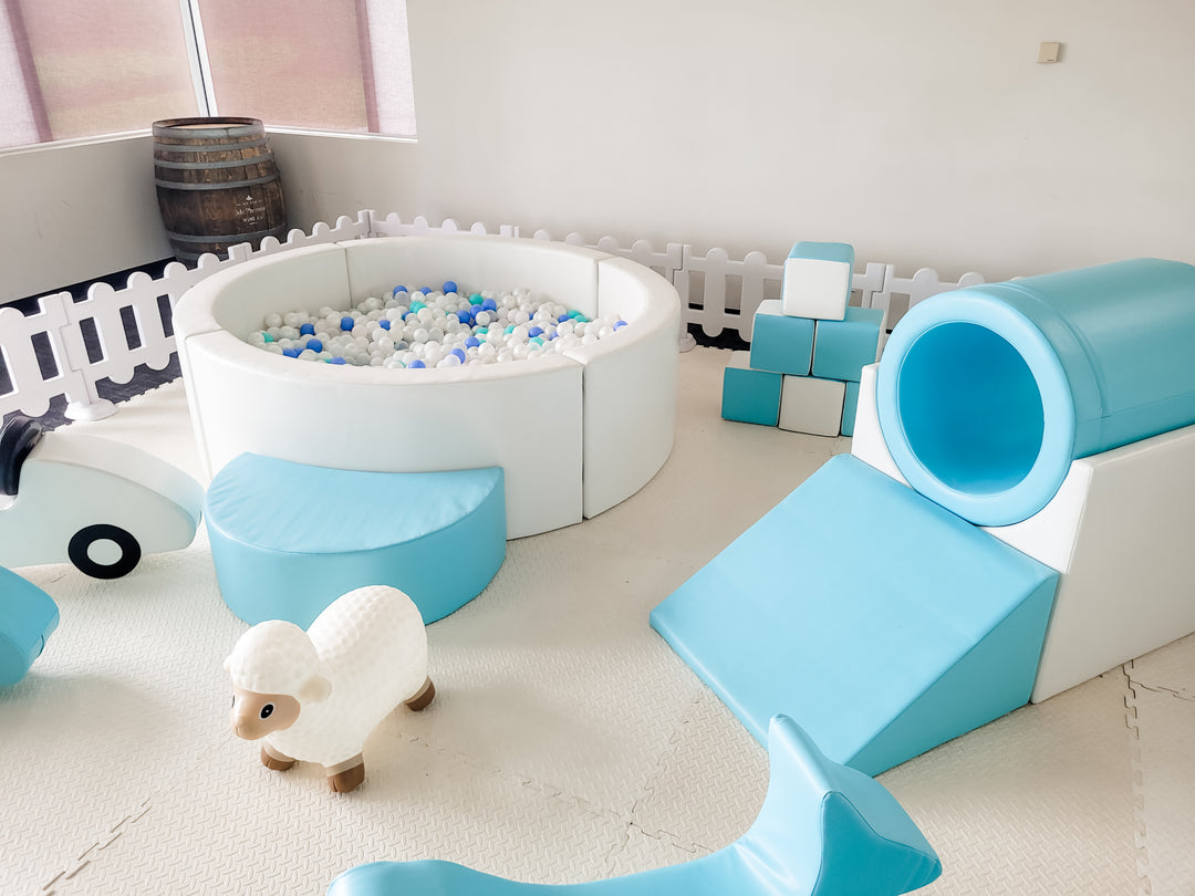 White ball pit with blue and white balls, baby blue tunnel climber, slide and blocks and a white sheep ride-on toy inside a fence