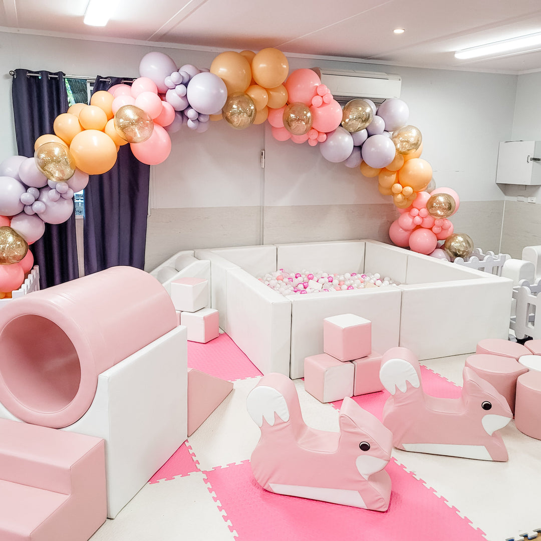 Baby pink ring tunnel, blocks and two bunny rockers by a square white ball pit under a peach, lilac, pink and gold garland