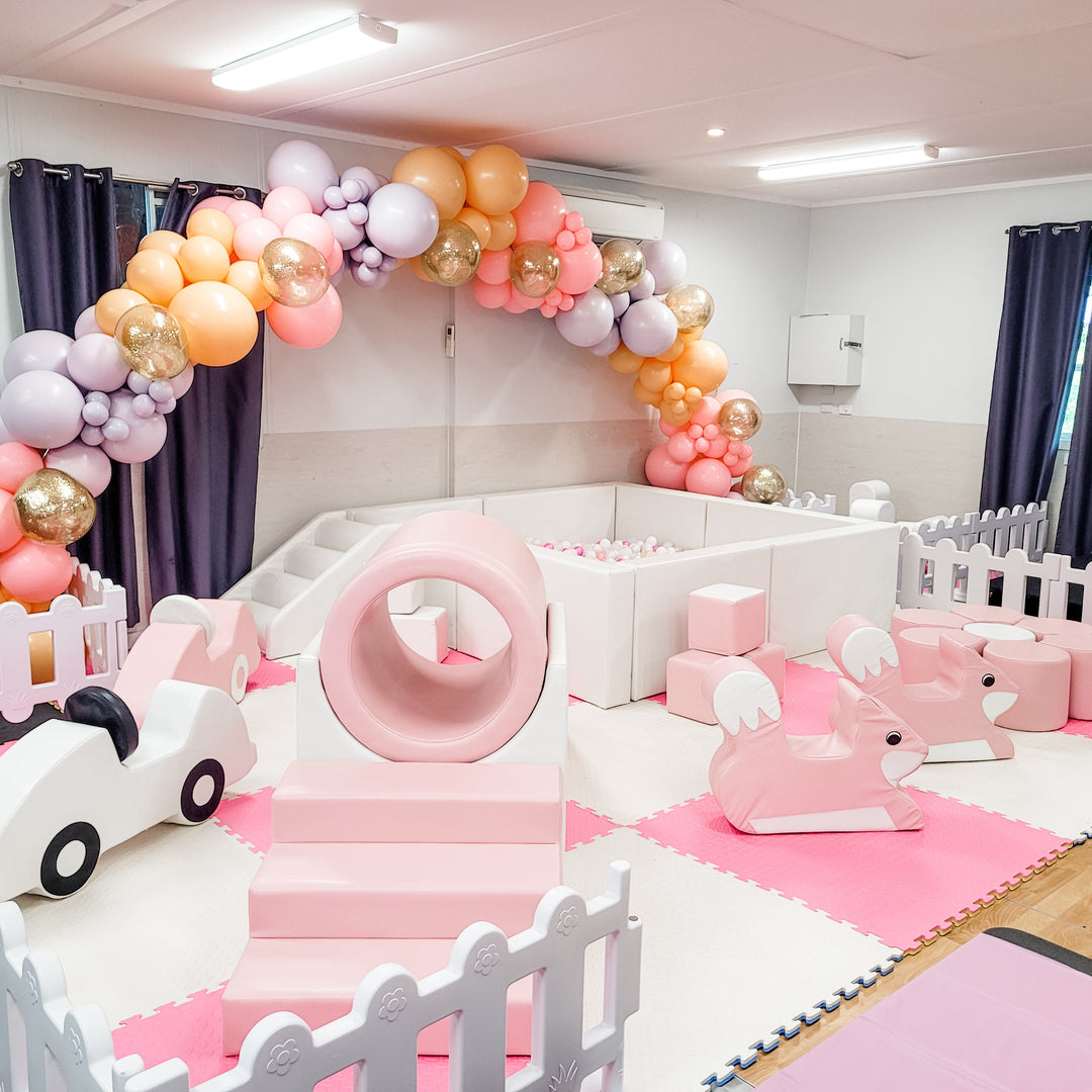 Wide view of the pink soft play with a white car, steps, ring tunnel, square ball pit and bunny rockers under a pastel garland