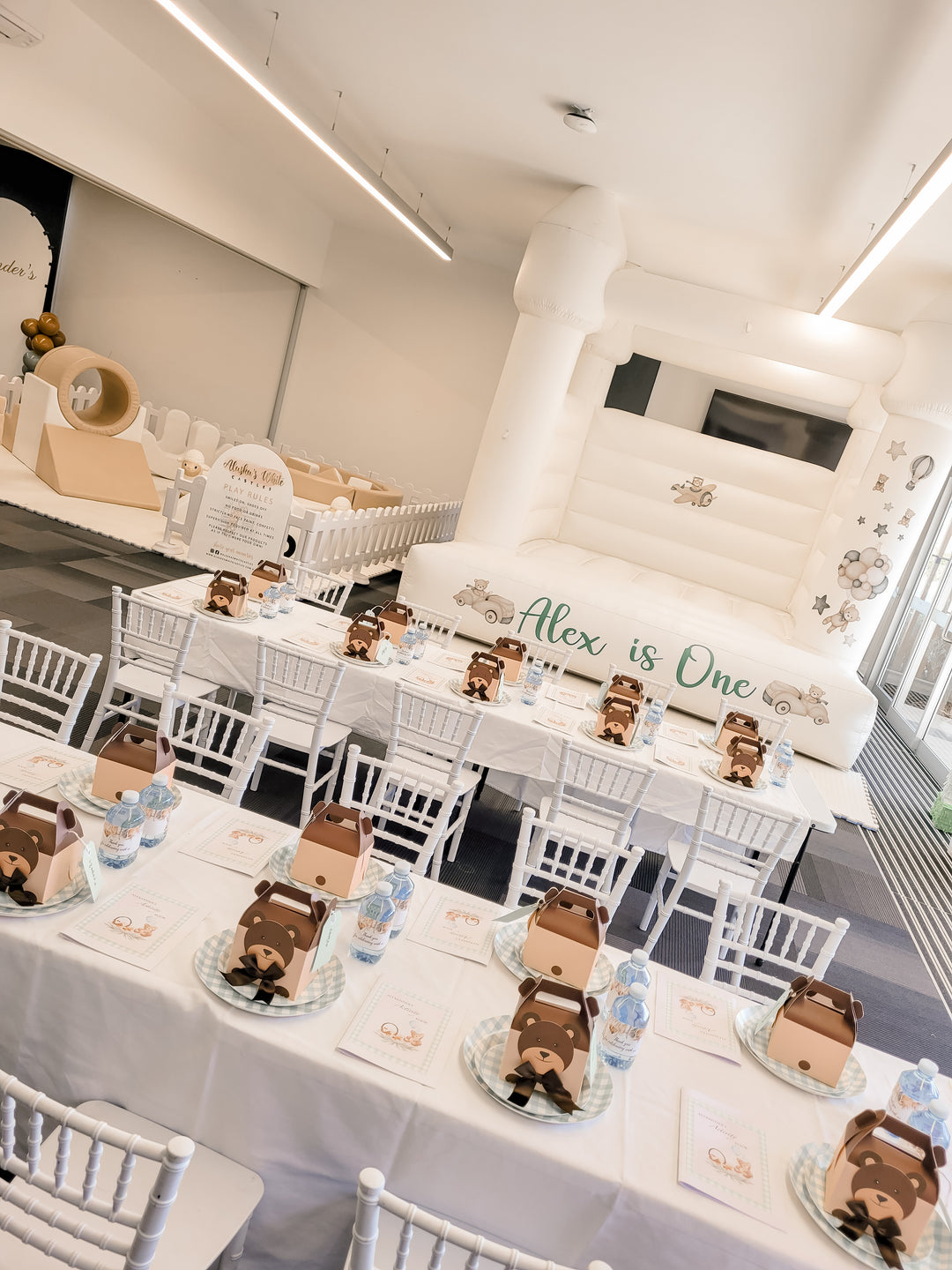 Close-up of teddy bear favour boxes on kids tables with white chairs in front of the Alex is One white bouncy castle