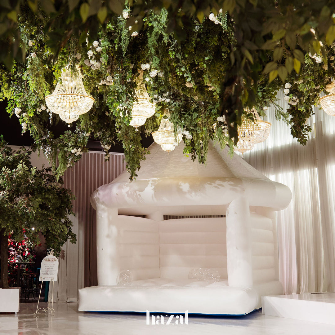 Wide view of the Dome white wedding bouncy castle with clear bubble balloons inside, framed by greenery and chandeliers