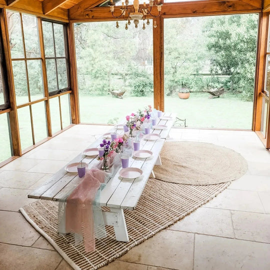 Low boho rustic trestle table with lilac cups, pink plates and flowers on jute rugs in a sunroom for party hire in Melbourne