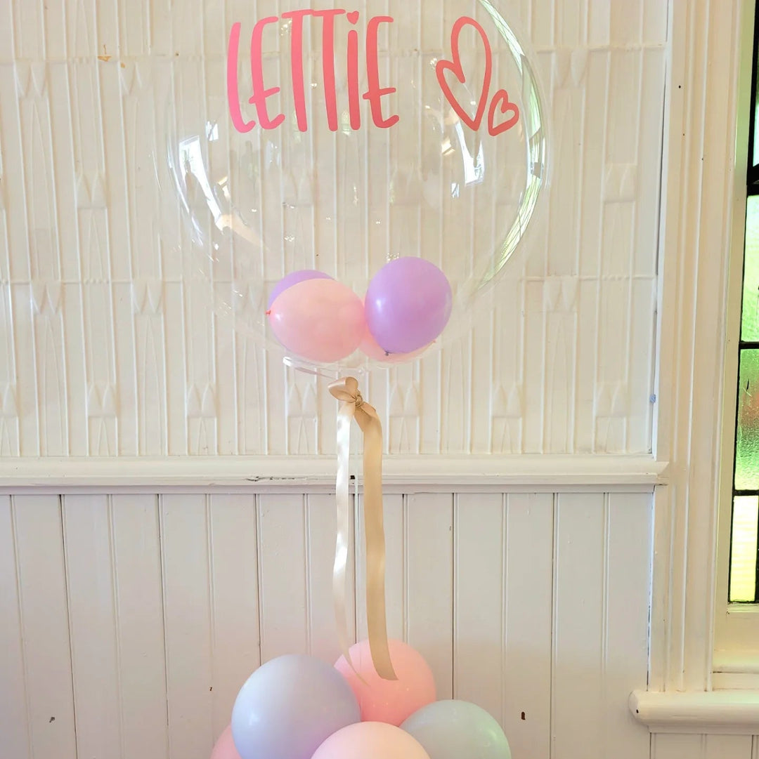 Clear bubble balloon with a pink Lettie name decal and heart, filled with mini pink and lilac balloons above a pastel cluster